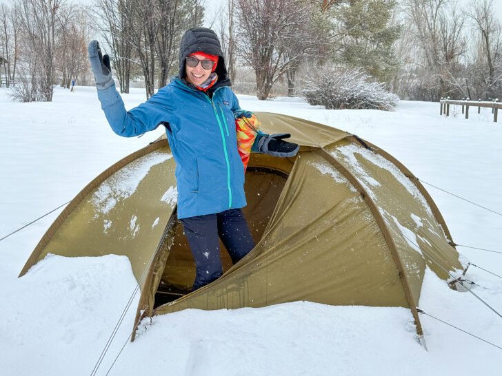 A woman pops out of a Fjallraven tent with snow piled up around it.