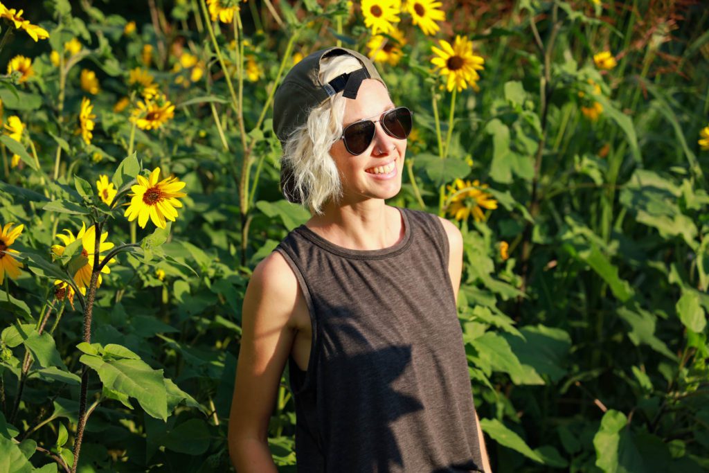 A woman smiles in front of sunflowers. She is wearing Sunski Sunglasses.