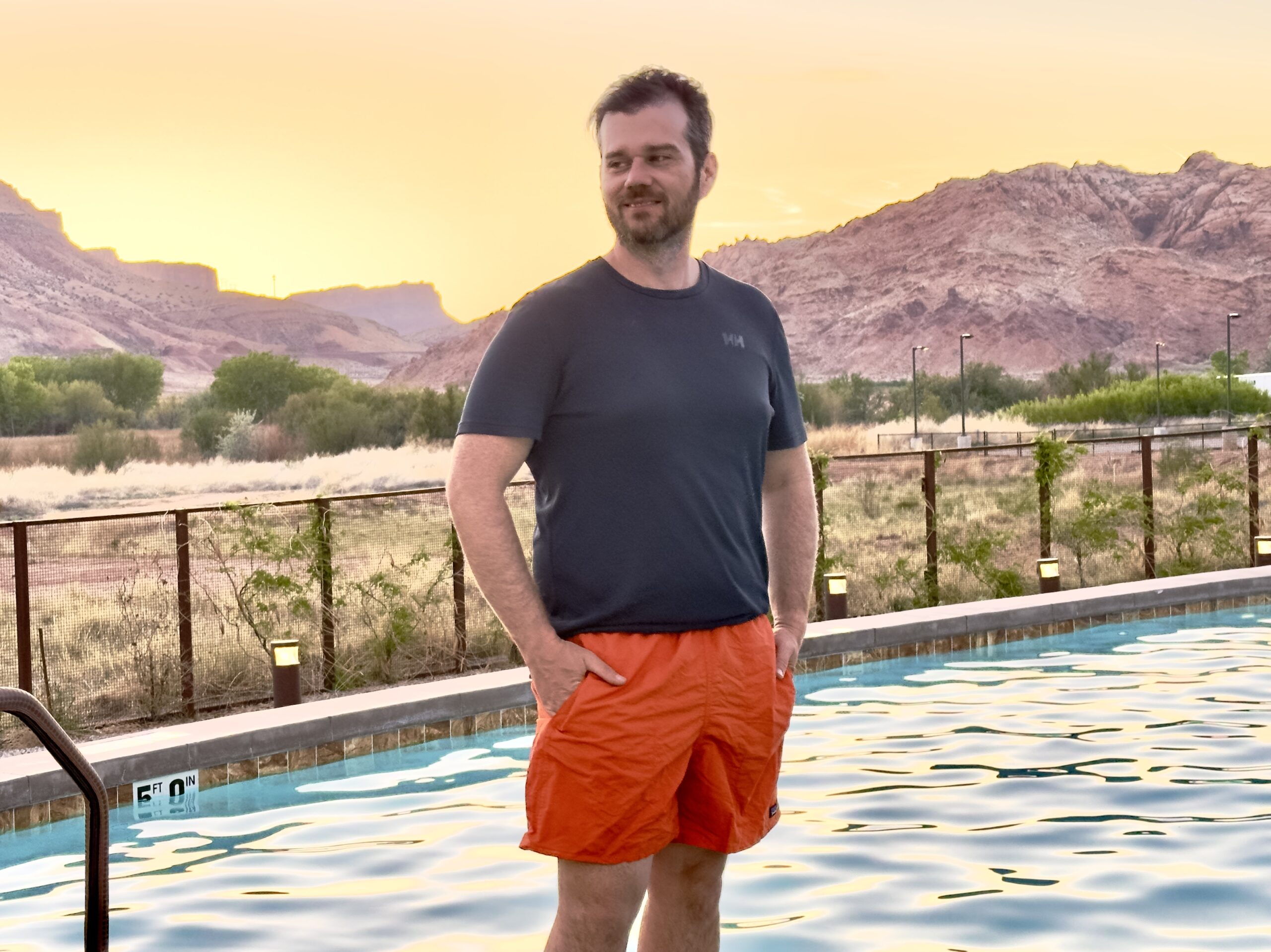 A man stands next to a pool in the desert in a blue tech tee and orange Patagonia Baggies sustainable swimwear for men.
