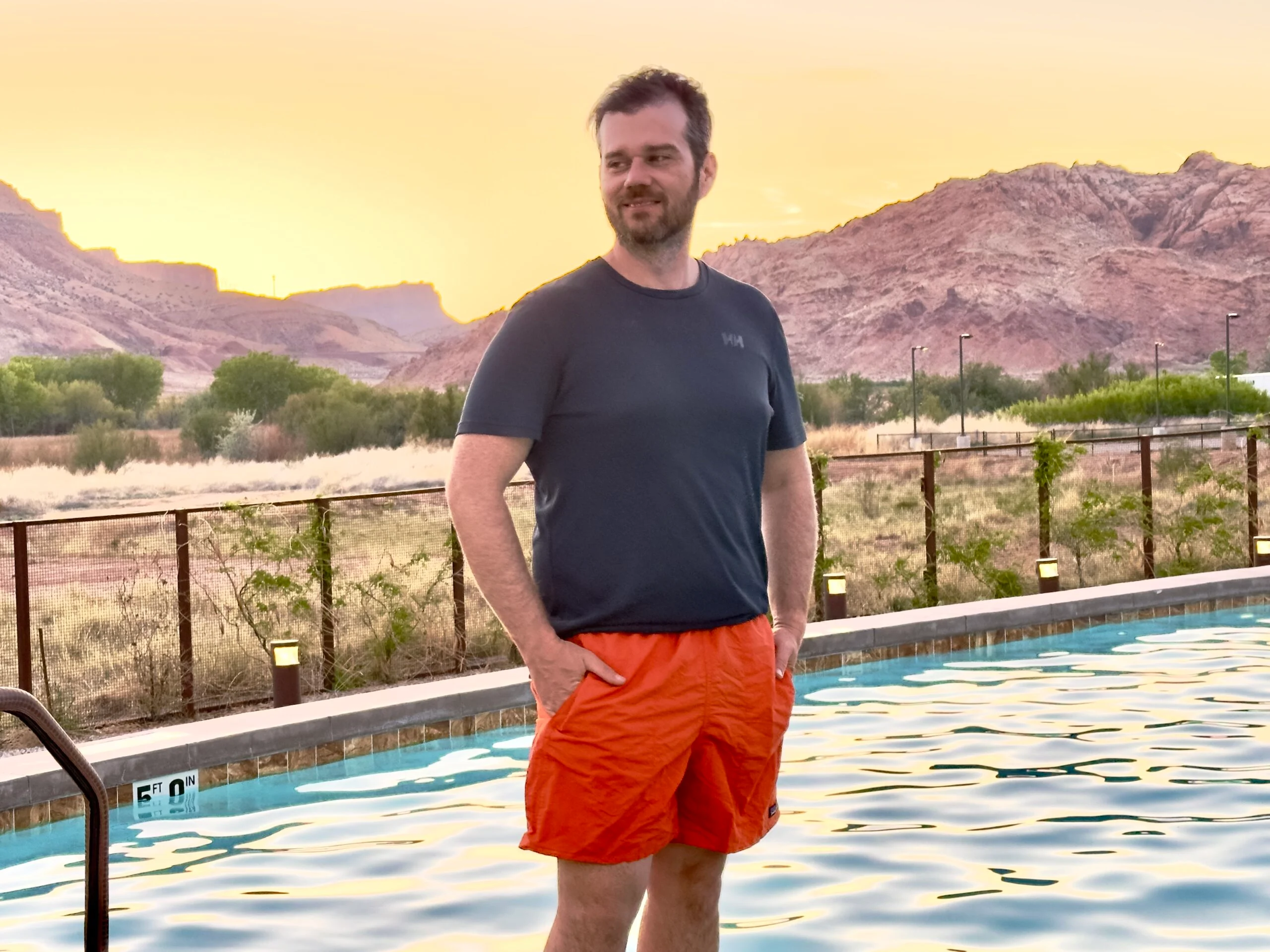 A man stands next to a pool in the desert in a blue tech tee and orange Patagonia Baggies sustainable swimwear for men.