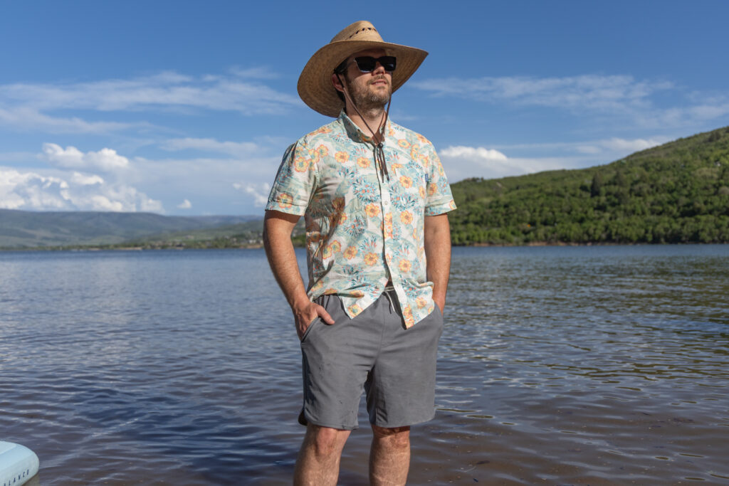 A man stands in the water in a large sun hat, Hawaiian shirt and Free Fly Apparel Reverb shorts.