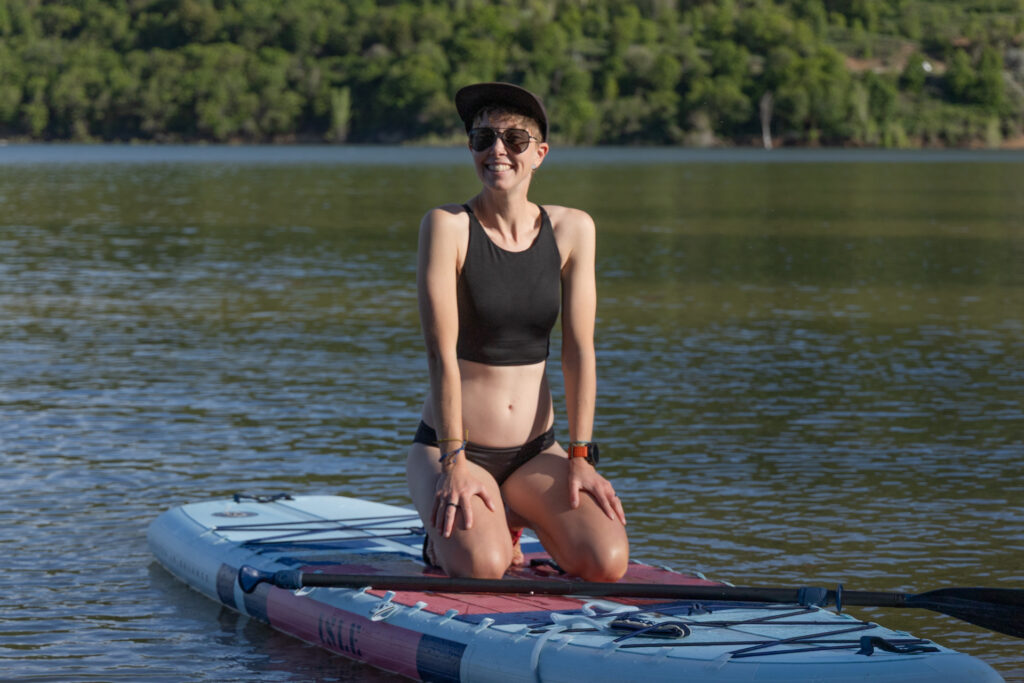 A woman kneels on a paddle board wearing the Patagonia Cami sustainable swimsuit top and Sunamee sustainable swimsuit bottoms.