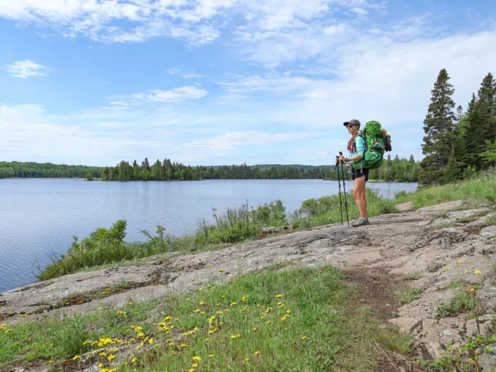 Woman stands on the edge of a lake with a backpack and trekking poles in Isle Royale National Park.
