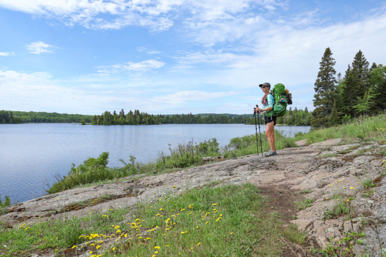 Woman stands on the edge of a lake with a backpack and trekking poles in Isle Royale National Park.