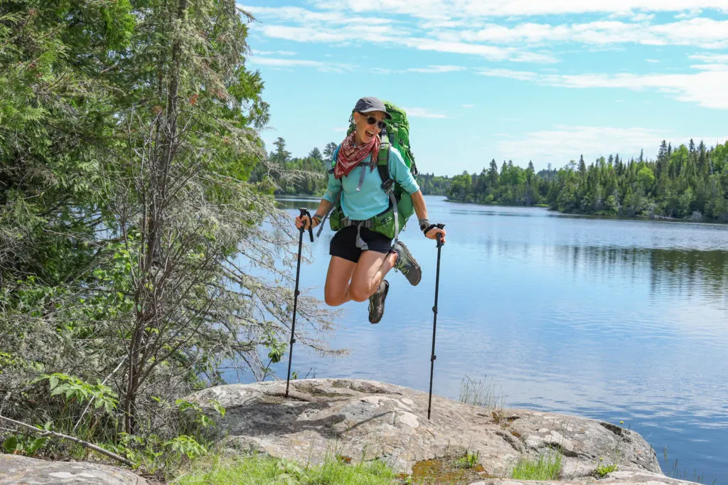 A woman jumping for joy in front of a lake with a backpack on.