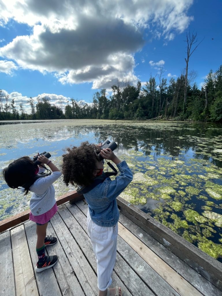 Photo by Jenna Tittelfitz: children peering through binoculars over a lake. 