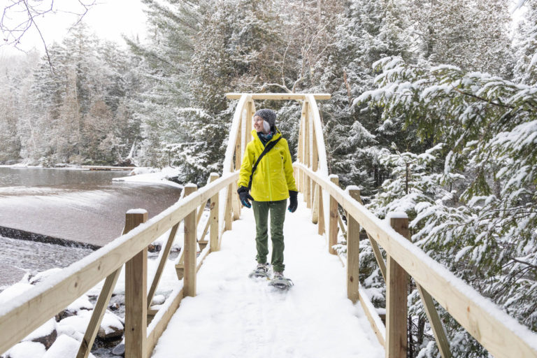 woman walking over a footbridge in the snow with snowshoes.