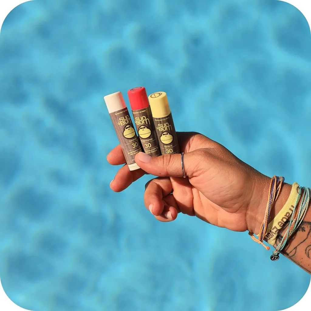 A hand holding three tubes of Sun Bum vegan lip balm over a swimming pool (photo courtesy of Sun Bum).