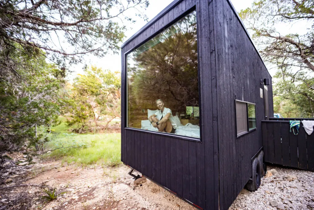 A woman smiles out the large window of a Getaway tiny cabin in Wimberley, Texas.