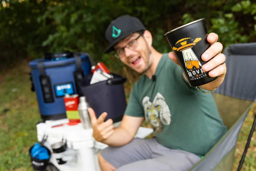 A man smiles outdoors and holds out a metal cup containing a cocktail.
