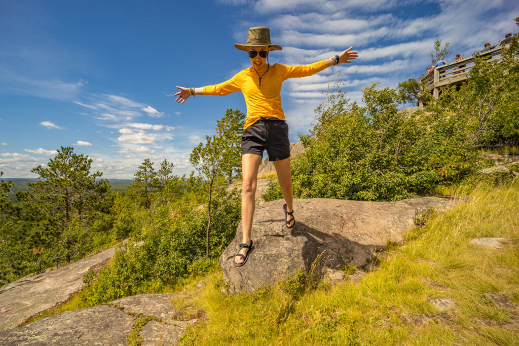 A woman jumps off a rock in the mountains while wearing hiking sandals.