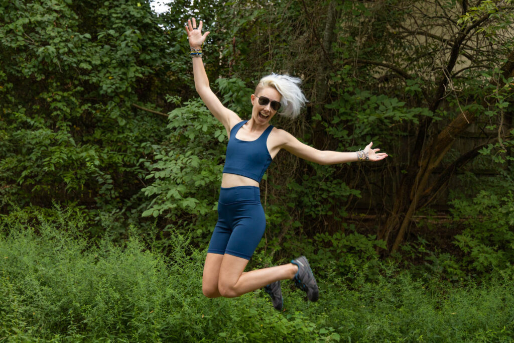 A woman leaps for joy outdoors in the tentree InMotion Longline Bra and Bike Short. 