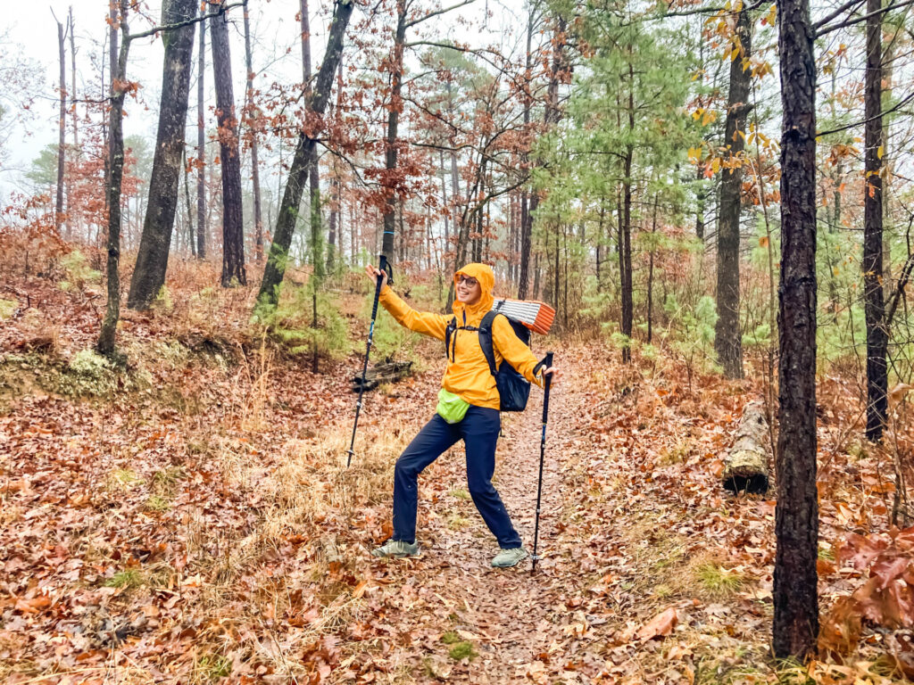 A woman in a rain jacket and backpack poses on a hiking trail.