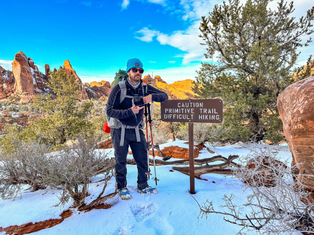 A backpacker poses next to a sign on a snowy trail that reads "Caution: Primitive trail difficult hiking." He is wearing micro spikes on his boots.