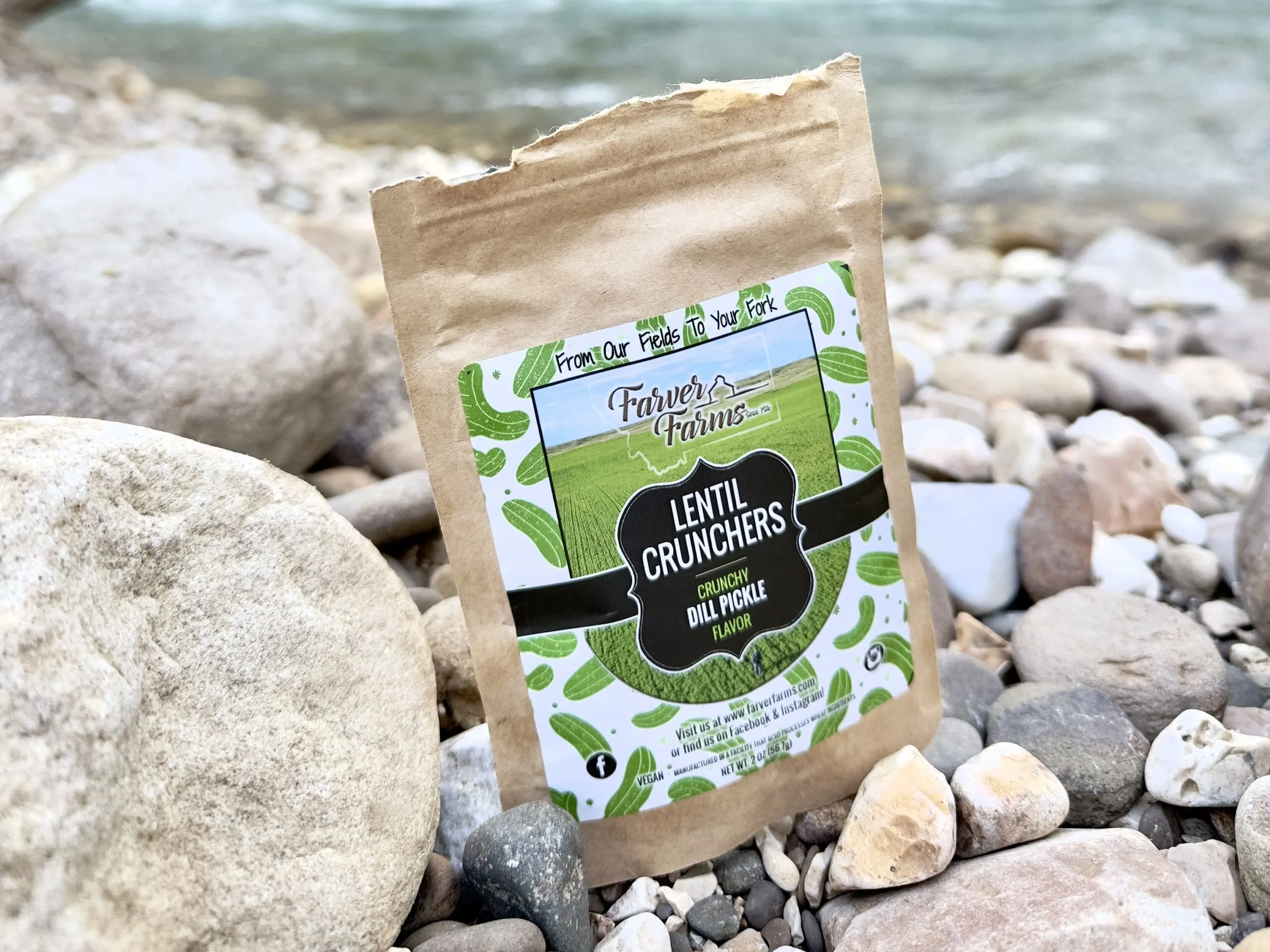 a 2 oz. sample pack of Farver Farms Lentil Crunchers in dill pickle flavor sitting on rocks by a river.