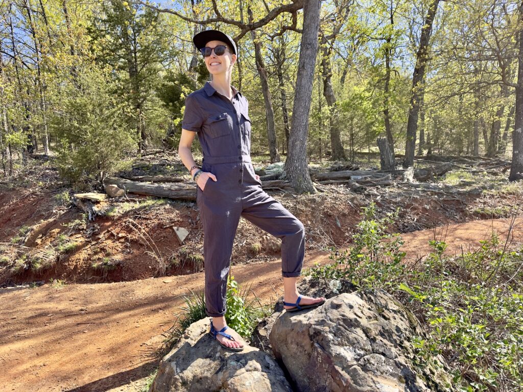 A woman stands on a rock next to a hiking trail wearing barefoot sandals. She is smiling.