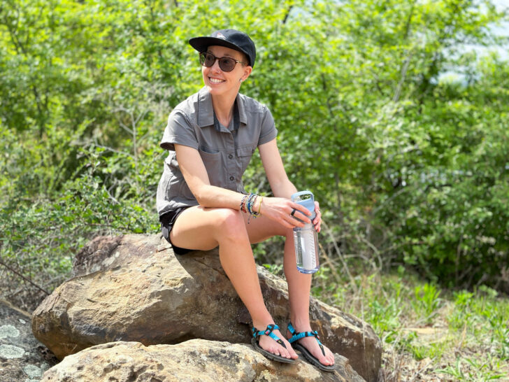 A woman sits on a rock smiling with a water bottle in hand.