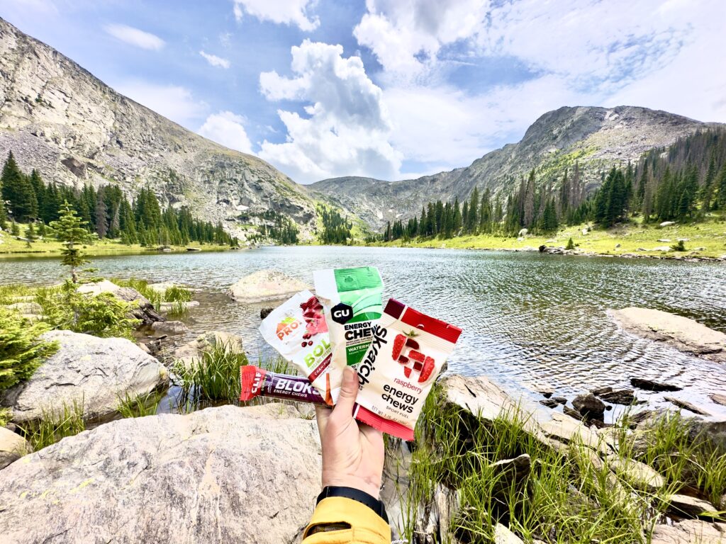A hand holds four packs of different vegan energy gummies up in front of a lake and mountains.