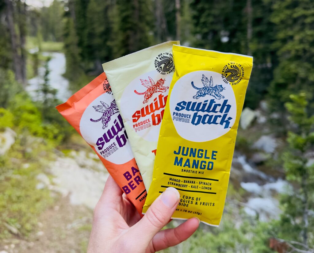 Three Switchback smoothie packets help up in front of treea and a river.
