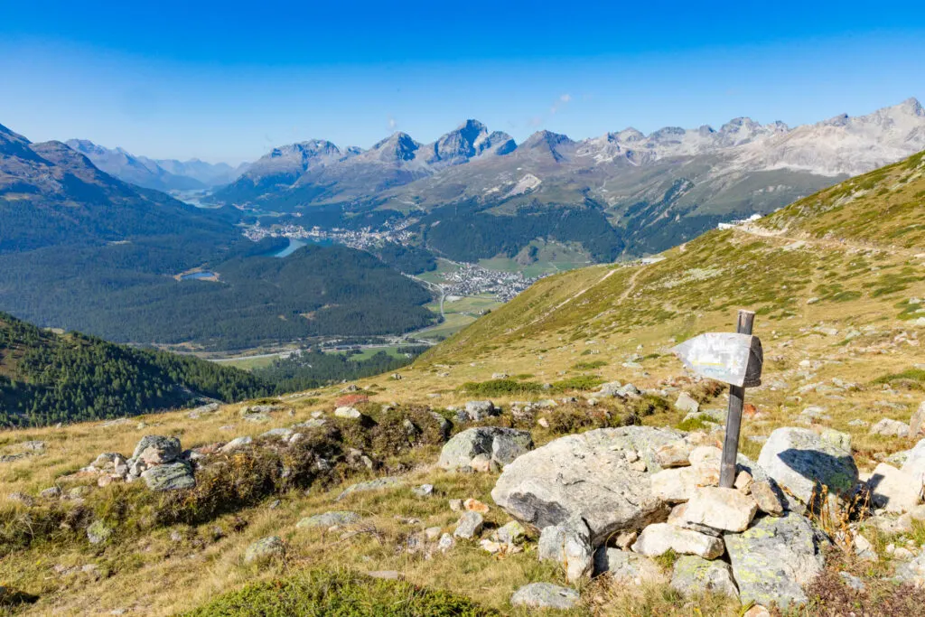 A view of mountains in the Swiss Alps, towns in the valley below, lakes and a signpost from the Panorama Trail in the Engadin.