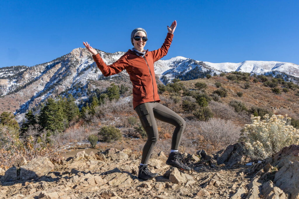 A woman poses happily in front of a mountain in hiking tights and a jacket.