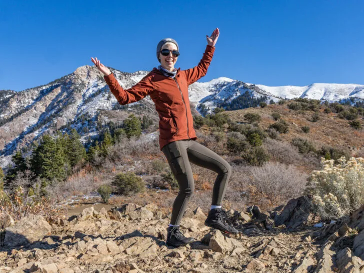 A woman poses happily in front of a mountain in hiking tights and a jacket.