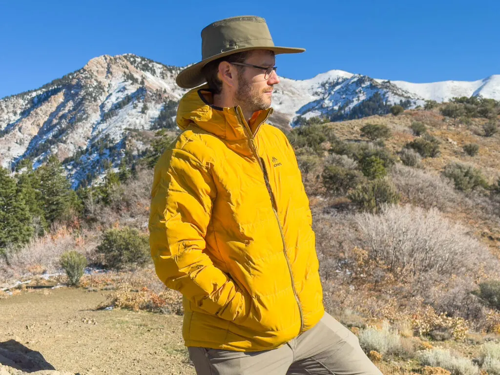 A man in the yellow Jones Dark Start Recycled Hoodie standing in front of snowy mountains.