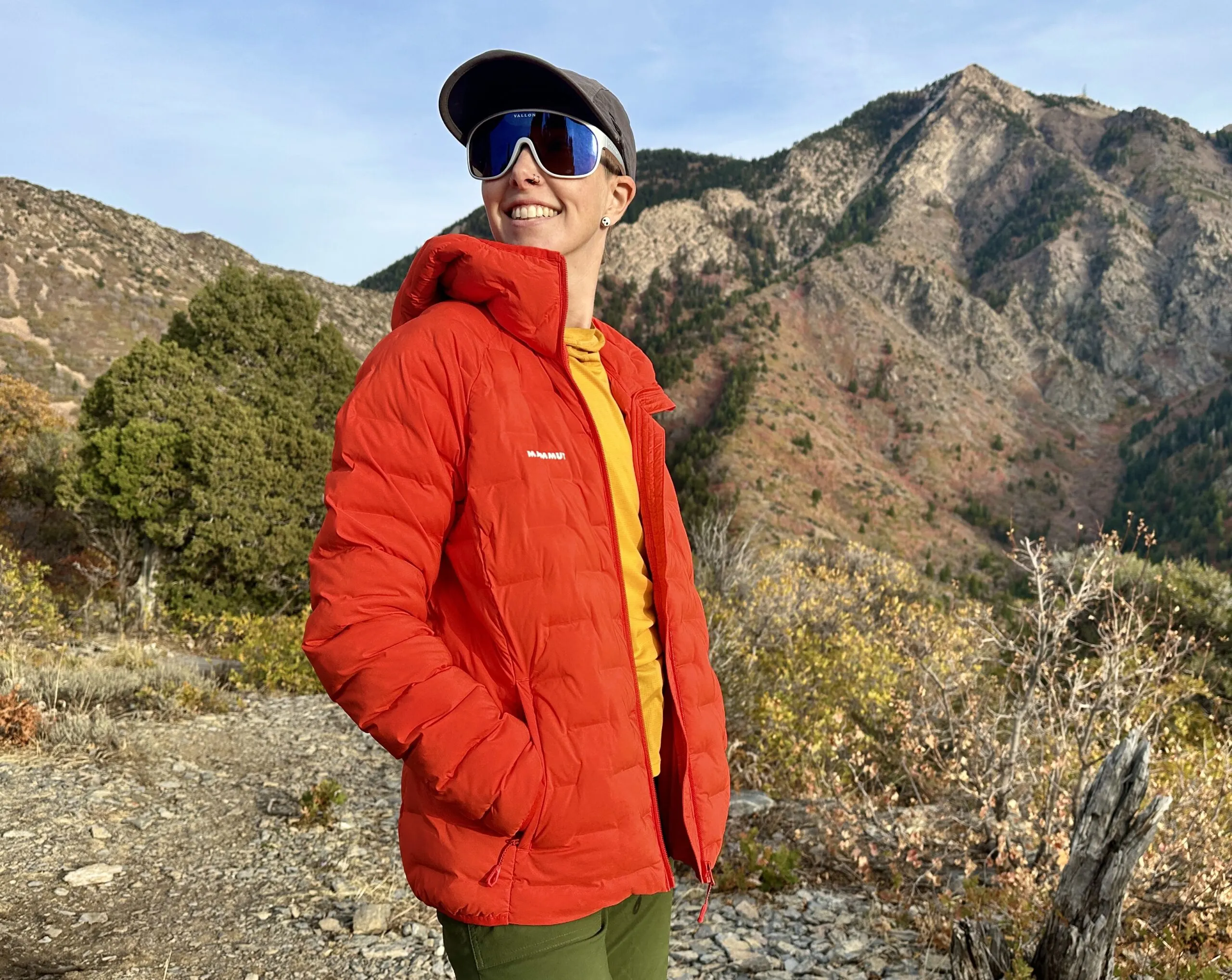 A woman smiles in front of a mountain wearing the Mammut Sender IN Hooded Jacket.