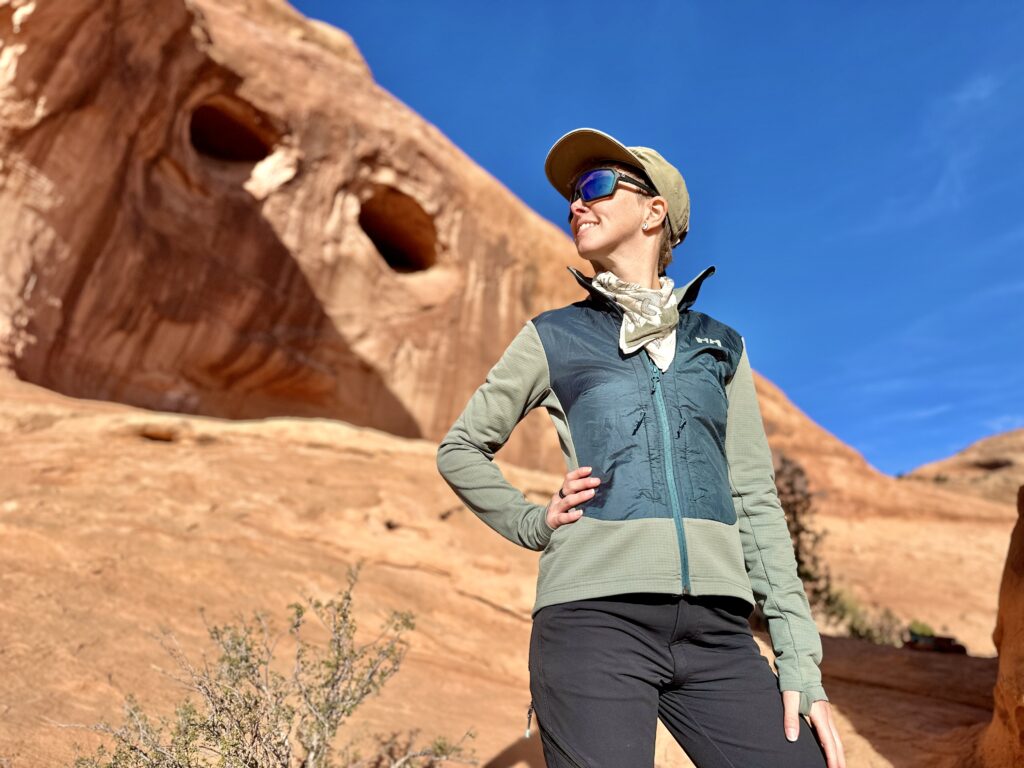 A woman stands smiling in front of red rocks wearing a Helly Hansen Versatile Hybrid Fleece Jacket.