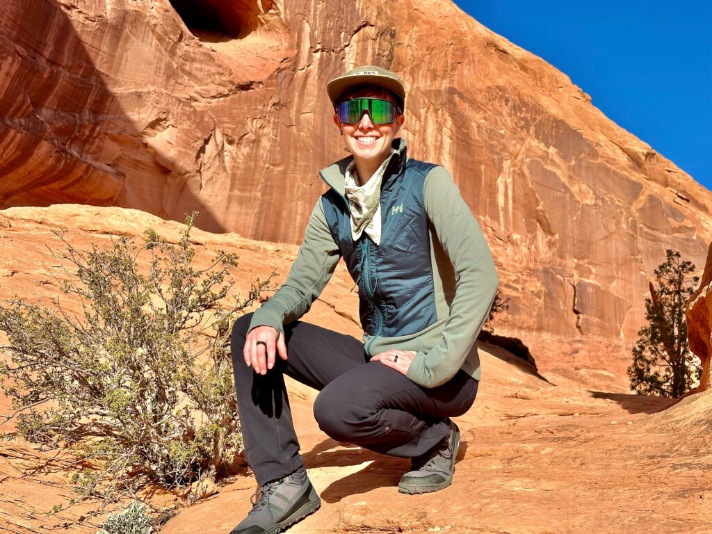 A woman kneels smiling in front of canyon walls wearing a Helly Hansen Versatile Hybrid Fleece Jacket.