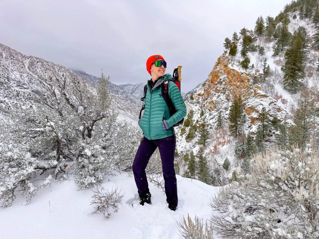 A woman in a hybrid jacket, sunglasses and a beanie stands in the snow in front of mountains with a backpack on.