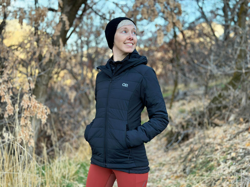 A woman in the woods smiles. She is wearing a beanie hat and the Outdoor Research Shadow Insulated Hoodie.