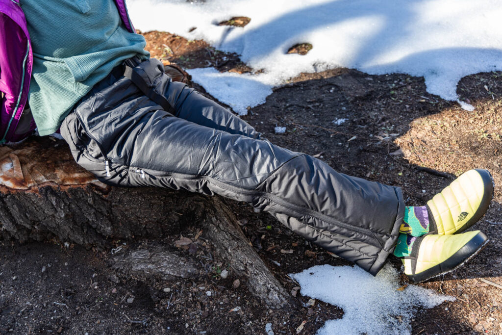 A woman sits on a stump next to snow wearing the Big Agnes Camp Boss insulated pants.