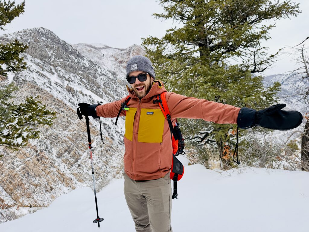 A man stands smiling in front of snowy mountains wearing a Patagonia R2 TechFace Hoody.
