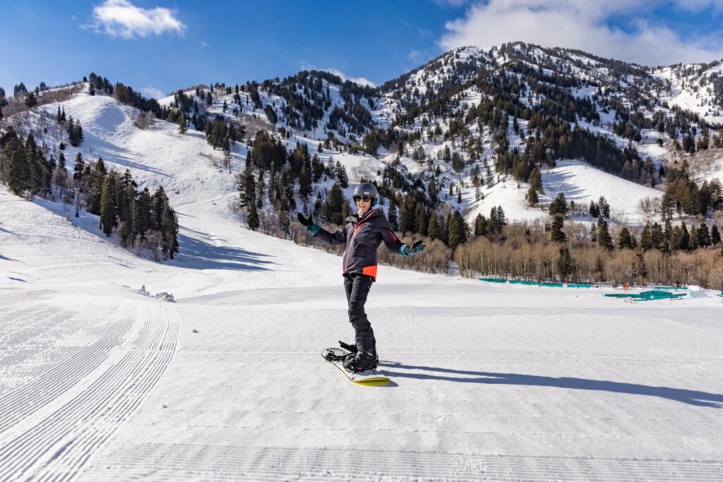 A woman smiles on a snowboard in front of mountains. She is wearing an insulated ski jacket.
