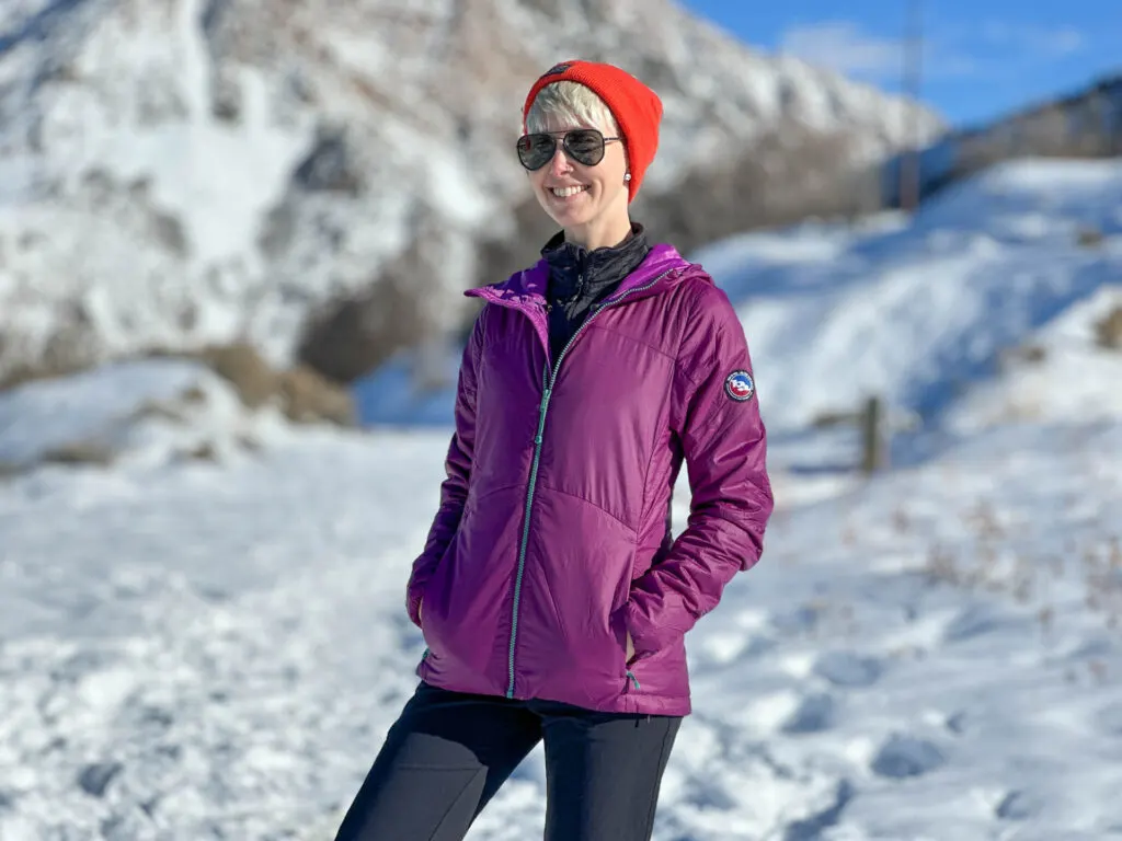 A woman smiles next to snowy mountains. She is wearing a Big Agnes Larkspur insulated jacket.