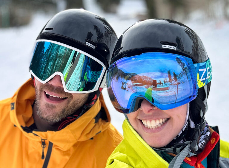 A man and woman smile. They are wearing ski goggles and helmets.