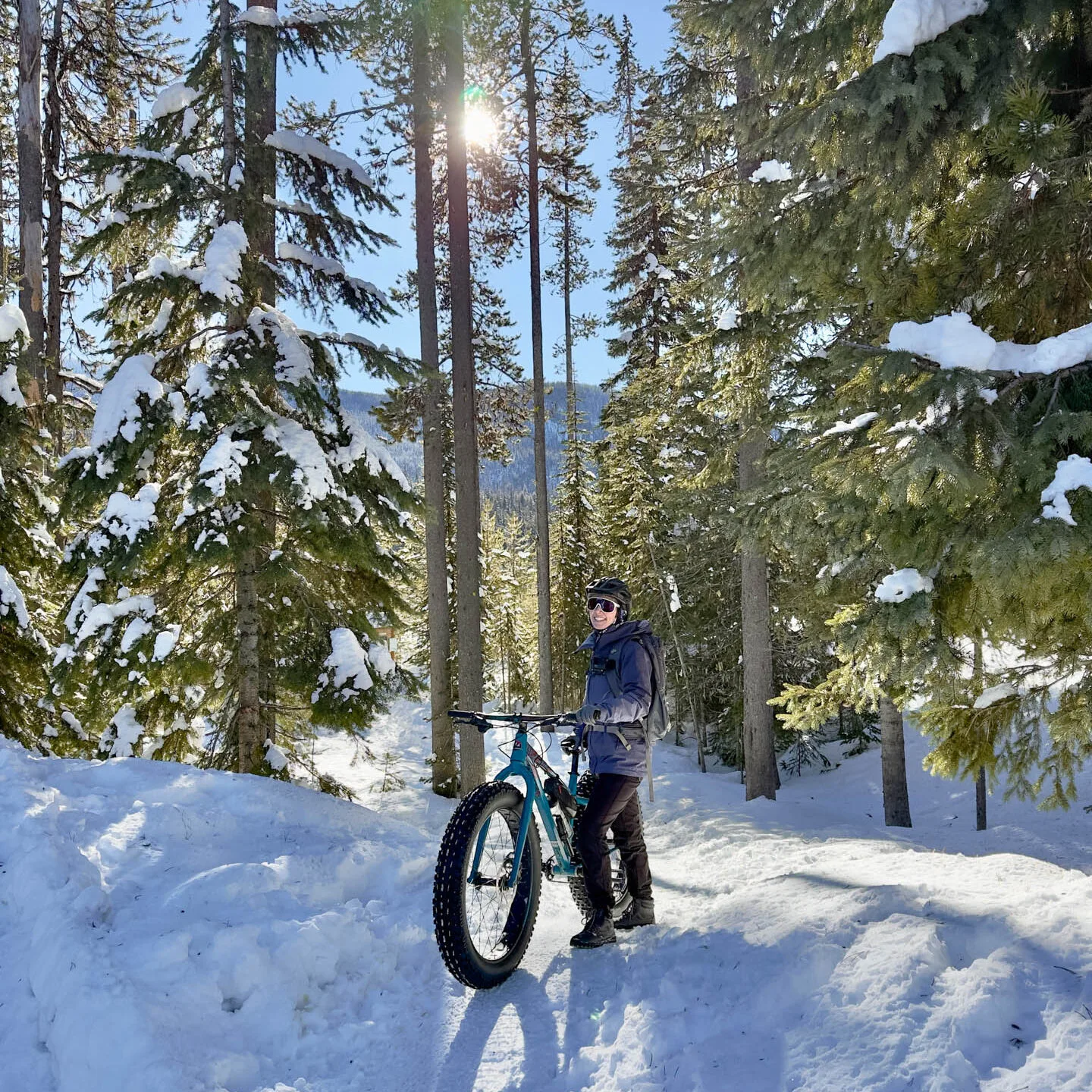 A woman stands smiling next to a fat tire bike on the snow. Snowy pines are behind her.