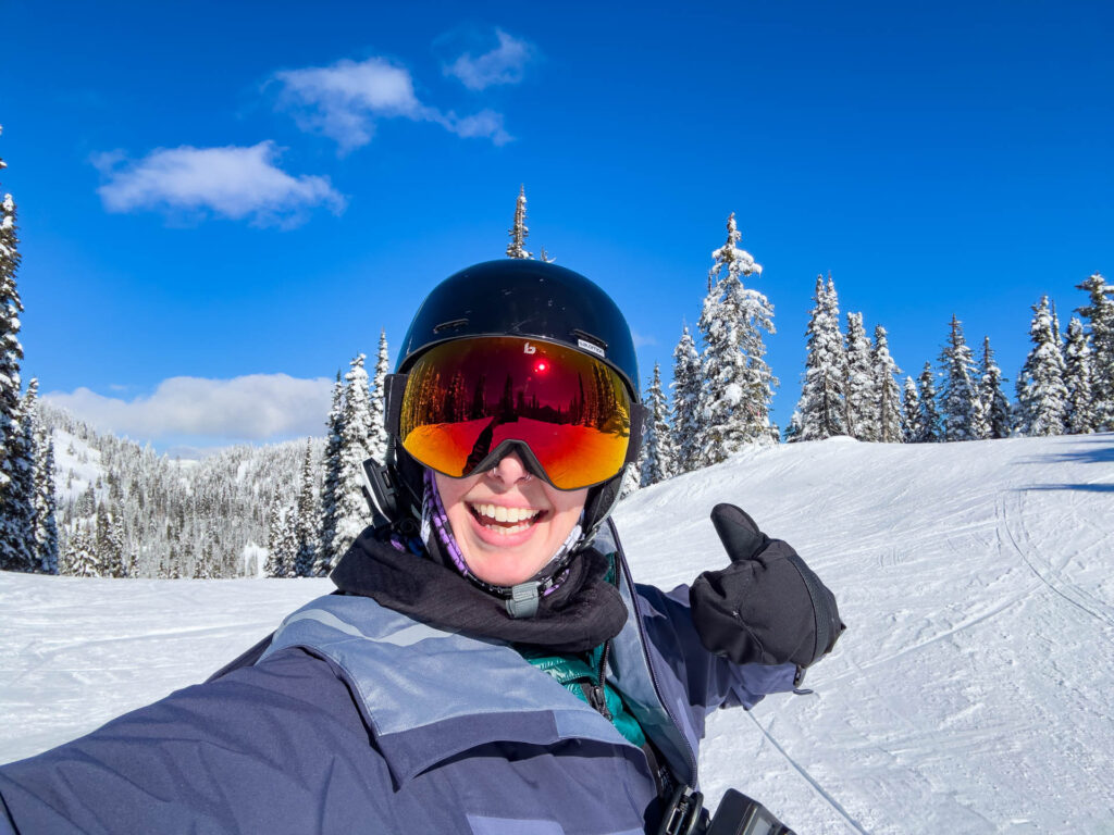A woman with ski goggles, a helmet and a Cardo Packtalk Outdoor device smiles. There are ski slopes and pine trees in the background.