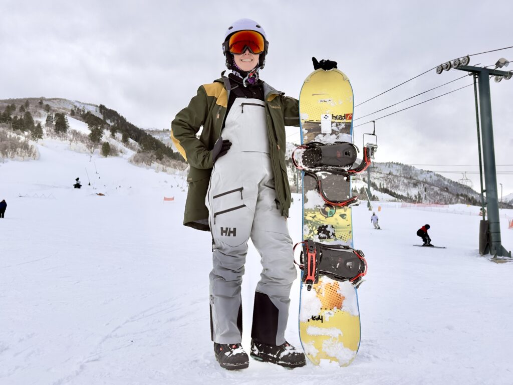 A woman stand with a snowboard in front of mountains. She is wearing a full ski kit.