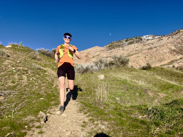 A smiling woman runs on a trail with mountains behind her.