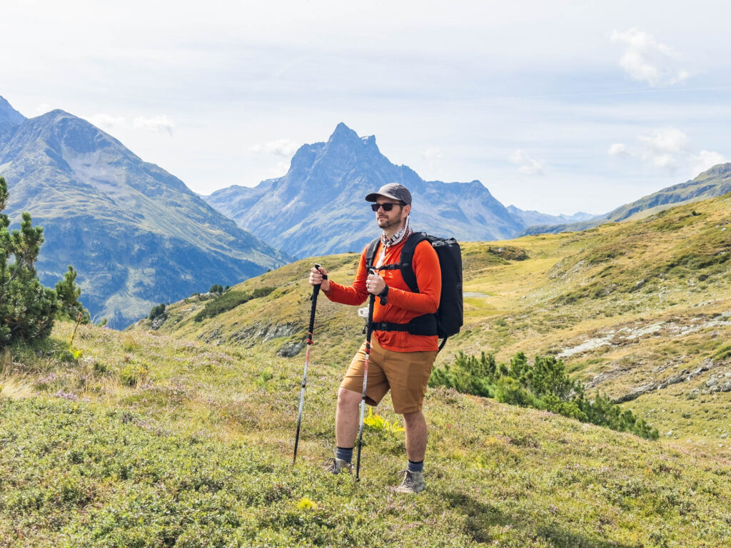 A man with a backpack and trekking poles stands in front of a mountain range.
