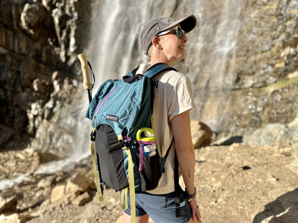A woman stands by a waterfall wearing a Gossamer Gear Loris 25 backpack.