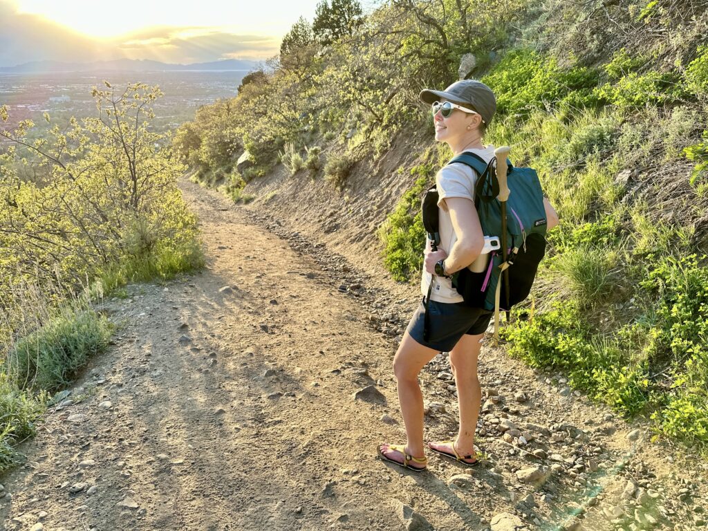A woman smiles from a rocky hiking trail. She has on a backpack and barefoot hiking sandals.