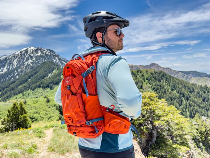 A man stands in front of a mountain with mountain bike apparel and an Osprey backpack.