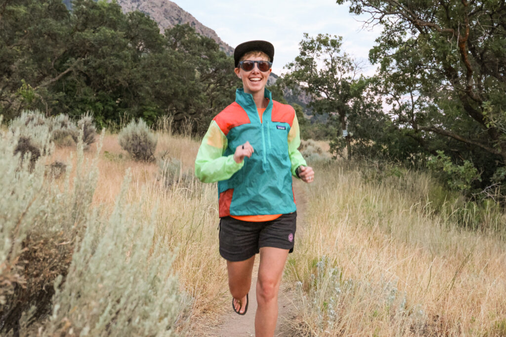 A woman smiles as she trail runs in a Patagonia Houdini Stash Half-Zip Windbreaker.