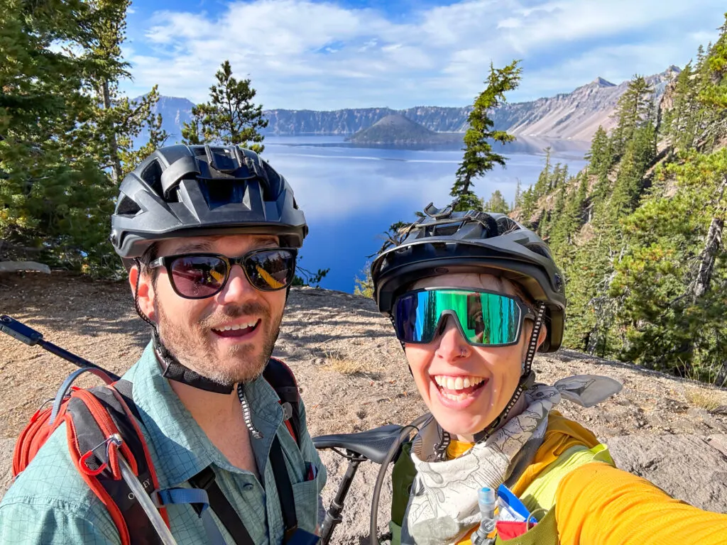 A couple with helmets smiling in front of an overlook with Wizard Island in the background during Ride the Rim at Crater Lake.