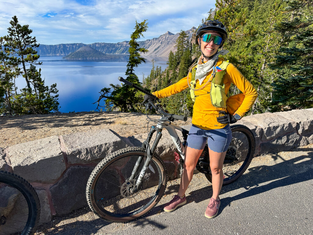 A woman with a bike smiling in front of an overlook with Wizard Island in the background during Ride the Rim at Crater Lake.