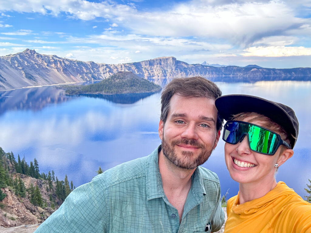 A couple smiling in front of an overlook with Wizard Island in the background at Crater Lake National Park.