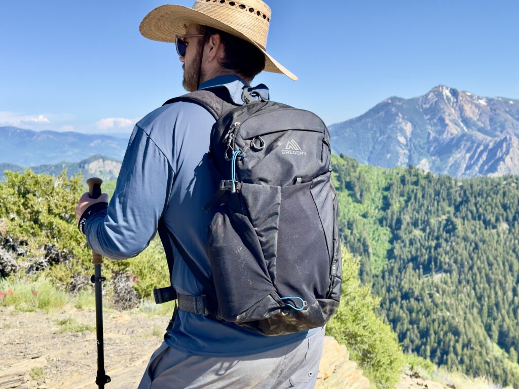 A man stands on a trail in front of mountains carrying a black Gregory Kiro daypack.